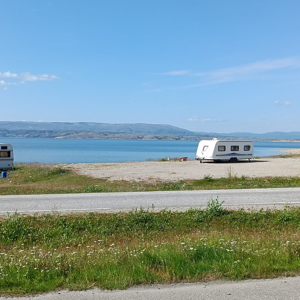 Grünstreifen dahinter Straße und Sandplatz. Darauf steht ein Wohnwagen. Dahinter ist das Meer zu sehen.